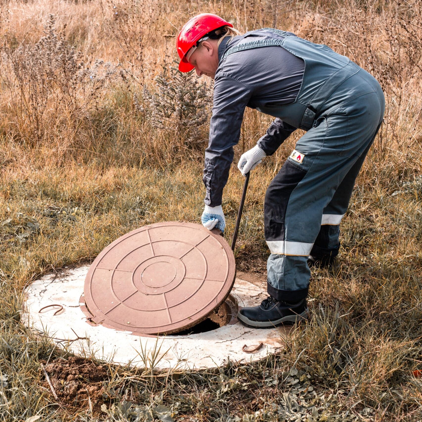 A worker in a hard hat lifts a manhole cover on a septic well. Inspection and maintenance of sewerage systems in rural areas